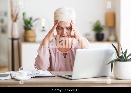 Sensazione di stanchezza e sottolineato. Senior business donna che lavorano su laptop, mantenendo gli occhi chiusi, toccando la sua testa, spazio di copia Foto Stock