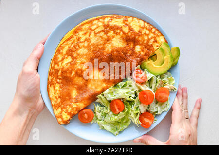 Uova fritte con verdure. le mani nel telaio. Frittata di vegetali su una piastra blu su un tavolo bianco, vista dall'alto Foto Stock