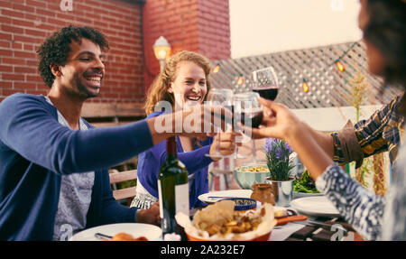 Millenial gli amici sorseggiando un bicchiere di vino rosso con cena al di fuori su di un patio sul tetto per il ringraziamento e godersi il tramonto Foto Stock