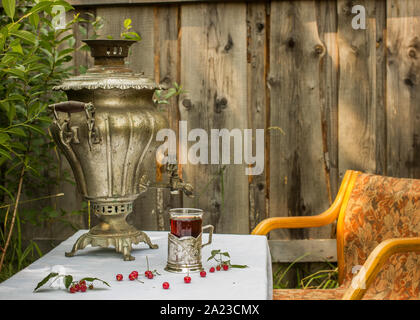 Vintage samovar in rame in un portabicchieri e un bicchiere di tè caldo sorge sulla tavola sono disseminate le ciliegie e lamponi, e vale la pena di sedia in RU Foto Stock