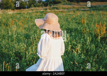 Splendida e romantica ragazza preteen in abito bianco e il cappello di paglia da dietro al prato, ora d'oro Foto Stock