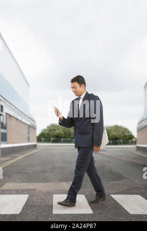 Imprenditore in abiti formali che trasportano borsa da ufficio attraversando la strada mentre utilizzando il suo telefono cellulare Foto Stock