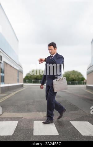 Imprenditore in abiti formali che trasportano borsa da ufficio guardando il suo orologio mentre attraversano la strada sulle strisce pedonali Foto Stock