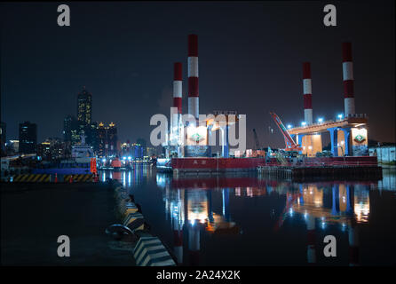 Kaohsiung, Taiwan: ciminiere e gru di un porto industriale / porto sito in Kaohsiung riflettendo in acqua durante la notte. Torre 85 in Background Foto Stock