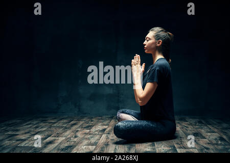 Vista posteriore della giovane donna a praticare yoga in seduta lotus posano con namaste nella stanza buia e copia di spazio. Ardha Padmasana esercizio , il concetto di meditazione Foto Stock