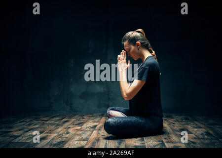 Vista posteriore della giovane donna a praticare yoga in seduta lotus posano con namaste nella stanza buia e copia di spazio. Ardha Padmasana esercizio , il concetto di meditazione Foto Stock