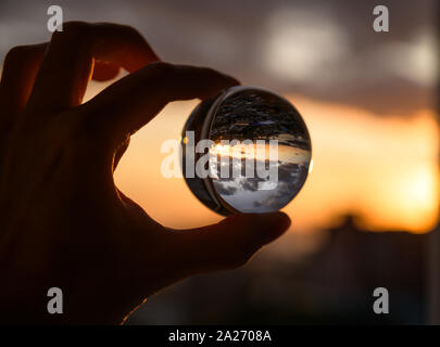 La mano trattiene Palla di vetro che riflette il Cielo di tramonto sulla città. Foto Stock