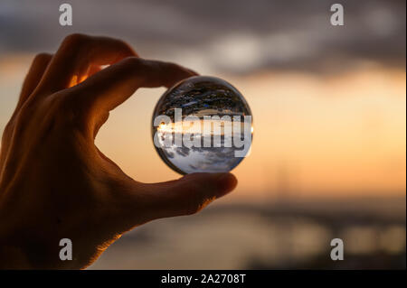 La mano trattiene Palla di vetro che riflette il Cielo di tramonto sulla città. Foto Stock