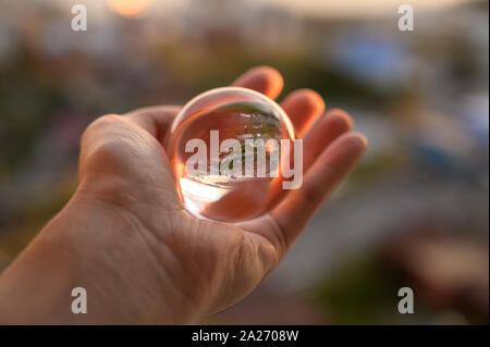 La mano trattiene Palla di vetro che riflette il Cielo di tramonto sulla città. Foto Stock