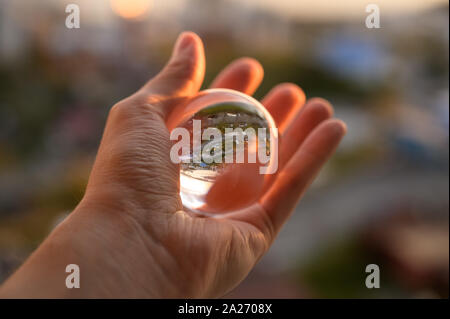 La mano trattiene Palla di vetro che riflette il Cielo di tramonto sulla città. Foto Stock