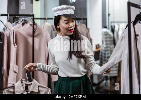 Bella giovane donna toccando diversi trench nel negozio di moda Foto Stock