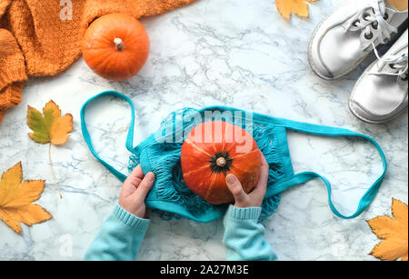 Autunno laici piatto con mani femminili mettendo arancione zucca in turchese stringa o borsa a rete. Vista dall'alto sulla luce Sfondo marmo bianco con scarpe, swea Foto Stock