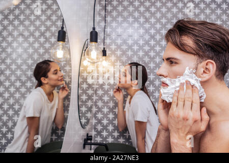 Uomo serio la rasatura e la sua fidanzata eliminando i suoi compongono nella loro stanza da bagno Foto Stock