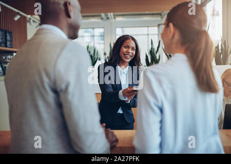 Sorridente giovane americano africano concierge in piedi dietro il banco della reception di un hotel per aiutare due clienti check in Foto Stock