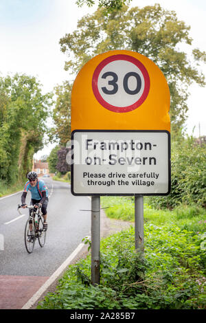 Un ciclista passando il traffico segnale di limite di velocità nel villaggio di Frampton-on-Severn, GLOUCESTERSHIRE REGNO UNITO Foto Stock