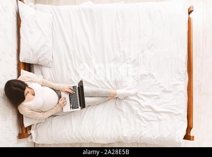 Donna incinta lavorando su laptop, seduta sul letto Foto Stock