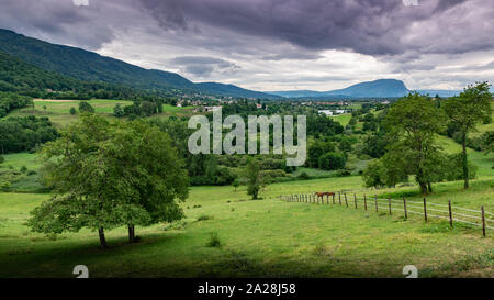 Paesaggio di montagne, foreste, piccola città, prati verdi e due cavalli su un pascolo recintato su uno sfondo di cielo nuvoloso.Machilly. Foto Stock