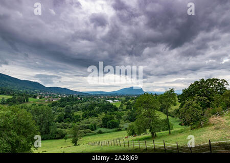 Paesaggio di montagne, foreste, piccola città, prati verdi e due cavalli su un pascolo recintato su uno sfondo di cielo nuvoloso.Machilly. Foto Stock