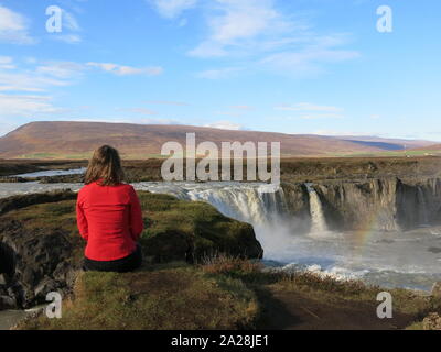 Una giovane donna si siede su una roccia e guarda fuori il vasto paesaggio vulcanico del Nord Islanda e le cascate Godafoss. Foto Stock