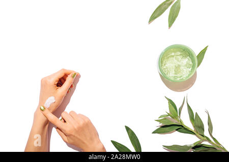 Mani femminili applicare crema idratante accanto a can di aloe gel e foglie verdi su sfondo bianco, piatto composizione laici. Foto Stock