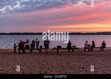 La gente seduta sulle panche in riva del lago a guardare il rosso e il blu del tramonto. Un gruppo di giovani si divertono alla spiaggia vicino lago. Party al tramonto. Foto Stock