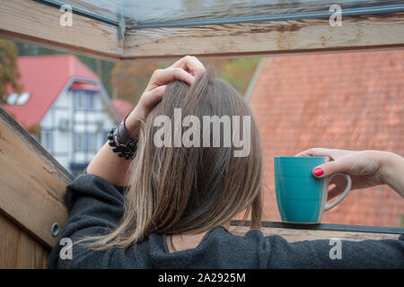 Donna o ragazza in piedi con una tazza di guardare attraverso la finestra a casa in un giorno di pioggia con una mano sulla testa Foto Stock
