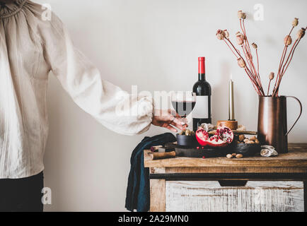Vino rosso, scheda di servire con formaggio, frutta, mandorle e olive, candele, decorativi fiori di cotone sul banco di cucina e womans mano azienda di vetro Foto Stock