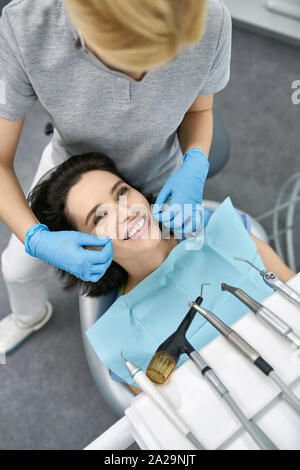 Bella ragazza in un paziente in bib una clinica dentale. Dentista in un grigio uniforme con maschera di protezione e guanti in lattice blu è il filo interdentale i suoi denti con Foto Stock