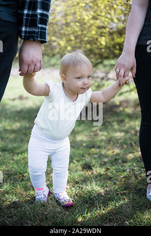 Un anno di età prendendo i primi passi, tenendo i suoi genitori le mani su una soleggiata giornata di primavera in un parco Foto Stock