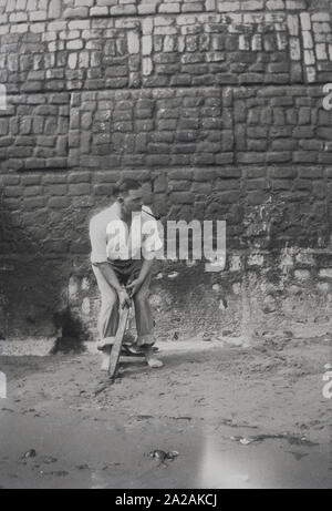 Anni '1930, storico, un uomo con pipa, pantaloni e maniche arrotolate, mazza in mano, giocando a cricket su una spiaggia, accanto a un muro alto, Inghilterra, Regno Unito. Foto Stock