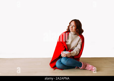 Sognare i giovani dai capelli rossi donna seduta sul pavimento isolato su sfondo bianco. Modello femminile sorridente guardando in alto, tenendo una tazza di caffè. Foto Stock