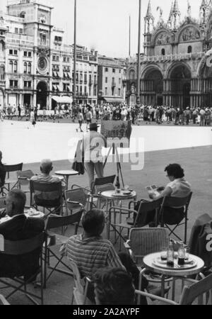 Sulla Piazza San Marco a Venezia, lasciato la Basilica di San Marco, la varietà dei passanti trambusto. Gli ospiti possono rilassarsi in un bar mentre la piazza stessa e gli edifici circostanti sono visitati da innumerevoli nativo e visitatori esotici. Un artista cattura la scena in un dipinto. Foto Stock