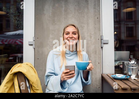 Incantevole ragazza bionda in attesa per il telefono messaggio mentre si beve il caffè sulla strada Foto Stock