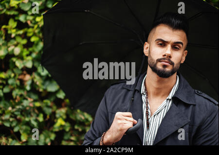 Alta moda barba arabo uomo usura sul mantello nero con ombrellone poste a pioggia meteo giorno. Foto Stock