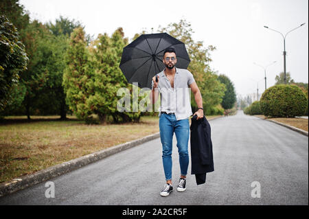 Alta moda barba arabo uomo usura sulla t-shirt, jeans e occhiali da sole passeggiate al parco con ombrellone e rivestire a portata di mano. Foto Stock