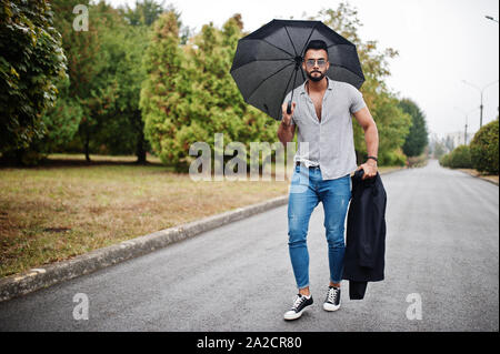 Alta moda barba arabo uomo usura sulla t-shirt, jeans e occhiali da sole passeggiate al parco con ombrellone e rivestire a portata di mano. Foto Stock