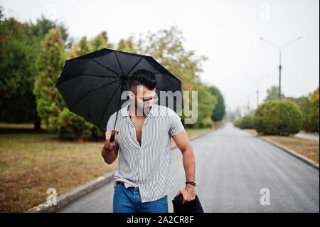 Alta moda barba arabo uomo usura sulla t-shirt, jeans e occhiali da sole passeggiate al parco con ombrellone e rivestire a portata di mano. Foto Stock