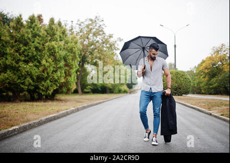 Alta moda barba arabo uomo usura sulla t-shirt, jeans e occhiali da sole passeggiate al parco con ombrellone e rivestire a portata di mano. Foto Stock