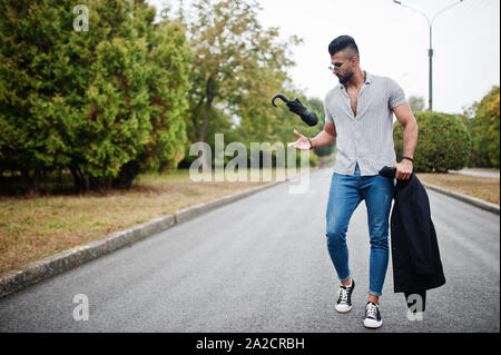 Alta moda barba arabo uomo usura sulla t-shirt, jeans e occhiali da sole passeggiate al parco con ombrellone e rivestire a portata di mano. Foto Stock