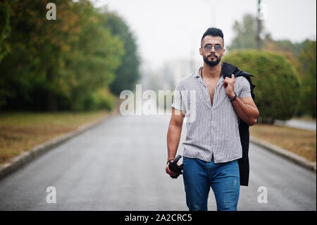 Alta moda barba arabo uomo usura sulla t-shirt, jeans e occhiali da sole passeggiate al parco con ombrellone e rivestire a portata di mano. Foto Stock