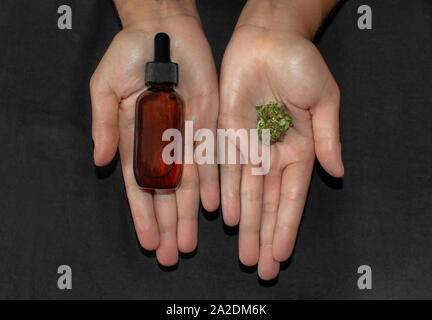 Che mostra le mani e il confronto di un bocciolo di marijuana e una bottiglia di olio in CBD, un potente estratto di cannabis. Rimedio naturale utilizzato per il dolore e le malattie. Foto Stock