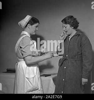 Eine junge Schwester des Deutschen Roten Kreuzes verarbreicht einer Kranken eine Tablette, Deutschland 1930er Jahre. Un giovane infermiere della Croce Rossa tedesca applicando una pillola per una donna malata, Germania 1930s. Foto Stock