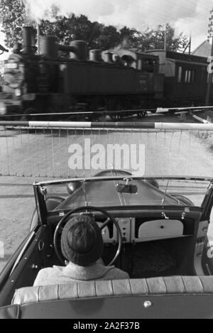 Eine junge Frau im Cabrio wartet auf das Vorbeifahren des Zuges un einer Schranke Donautal im, Deutschland 1930er Jahre. Una giovane donna alla guida di una cabriolet è attesa per il treno per passare da a un bar nel Danubio Dale, Germania 1930s. Foto Stock