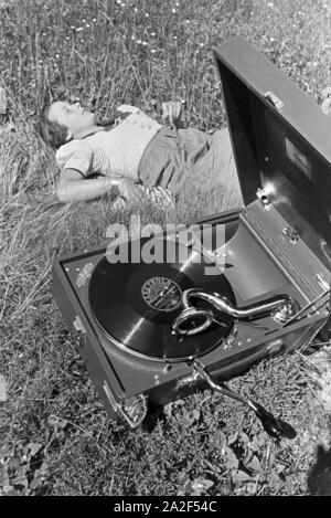 Eine junge Frau hört sich eine Platte der Schallplattenfirma Electrola mit dem Koffergrammophon auf der Wiese un, Deutschland 1930er Jahre. Una giovane donna di ascolto di un record dell'etichetta discografica Electrola giocato su un portatile wind-up gramophone, Germania 1930s. Foto Stock