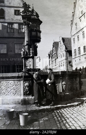 Zwei Frauen unterhalten sich während Sie die Wassereimer mit Wasser aus dem Springbrunnen vor dem imposanten Rathaus voll laufen lassen, Rothenburg ob der Tauber, Deutschland 1930er Jahre. Due donne aventi una chat mentre il bucktes sono il riempimento con acqua fuori della fontana posta di fronte all'imponente Municipio di Rothenburg ob der Tauber, Germania 1930s. Foto Stock