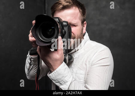 Un bel uomo - un fotografo in una camicia bianca spara in studio su una fotocamera reflex digitale, che si erge su un treppiede. Close-up. Foto Stock