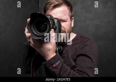 Un bel uomo - un fotografo in un maglione marrone spara in studio su una fotocamera reflex digitale, che si erge su un treppiede. Close-up. Foto Stock