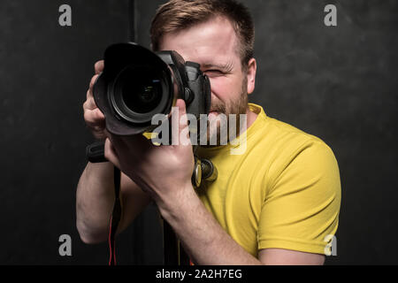 Un bel uomo - un fotografo in un giallo T-shirt spara in studio su una fotocamera reflex digitale, che si erge su un treppiede. Close-up. Foto Stock