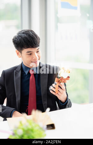 Un giovane imprenditore con un fiducioso suit seduti ad una scrivania in un ufficio moderno. Egli ha sorriso e guardando il trofeo d'oro a causa del successo aziendale. Foto Stock