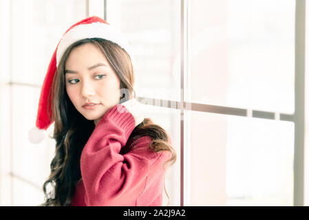 In prossimità di una giovane e bella donna asiatica che indossa un cappello da Babbo Natale è in piedi vicino alla finestra sul giorno di Natale, in attesa di un regalo a sorpresa dal suo boyfri Foto Stock